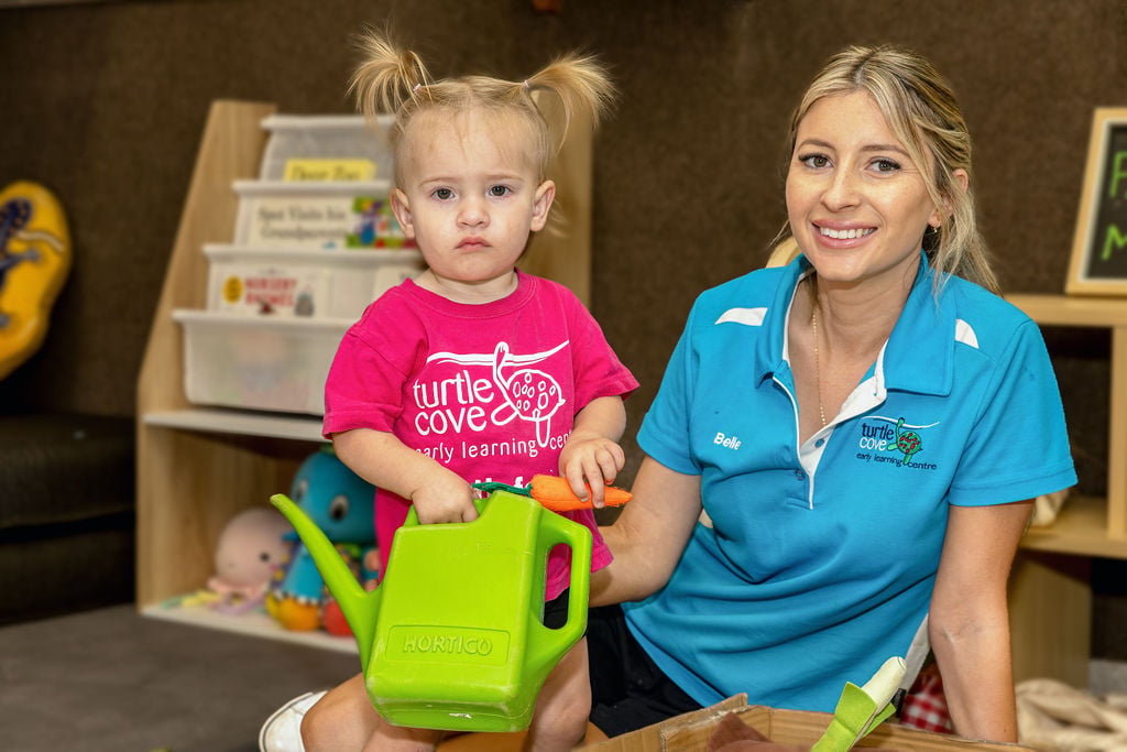 Toddler in pink t-shirt holding the watering can and toy carrot, and a teacher in turtle cover uniform smiling for the photo.
