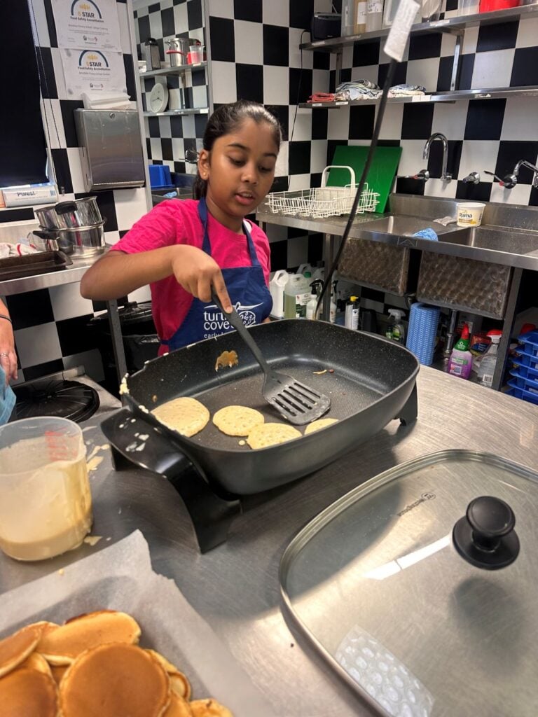 Child in a blue apron cooking pancakes on a grill pan during a kids cooking class